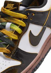 Nike-sneaker med en vit läderbas, svarta och orange detaljer, gröna och gula gradient snören, gummiyttersula och synliga sömmar.