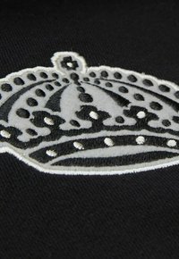 Schwarzer Stoff mit einem weißen gestickten Krondesign, das strukturierte Akzente und gepunktete Details entlang der Kontur der Krone aufweist.