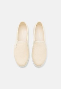 Beige slip-on sko laget av teksturert lerret, med elastiske sidepaneler og en hvit gummisåle. Minimalistisk design med subtil merkevare.