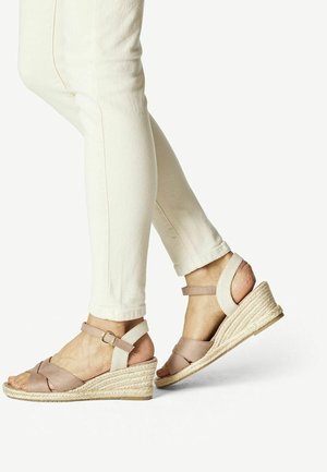 Jambes dans un pantalon ajusté écru portant des sandales compensées beiges avec des brides à la cheville et des talons en corde tressée sur un fond clair uni.
