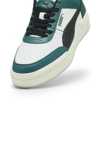 Puma tornacipő, zöldes-kék és fehér bőr felsőrészével, fekete részletekkel és texturált elemekkel. Lapos fekete fűzők és párnázott talp.