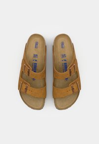 Bruna mockaskandskor med två justerbara remmar och metallspännen; anatomiskt utformad fotbädd; präglad varumärkeslogotyp; modern slip-on design.