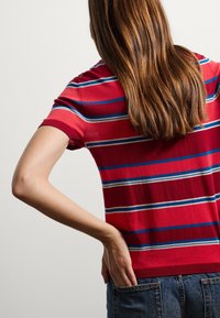 Person mit langen braunen Haaren, die ein rot gestreiftes Hemd und Jeans trägt und mit einer Hand ihren unteren Rücken berührt.