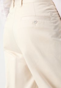 Pantalones color crema con una textura suave. Presenta un bolsillo trasero con un botón blanco y una sutil marca sobre el bolsillo.