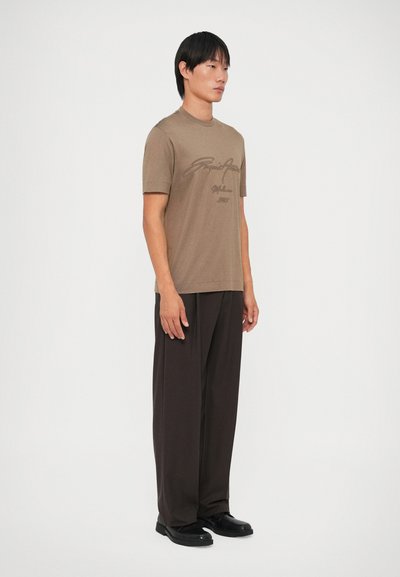 Camiseta marrón de manga corta con cuello redondo y estampado gráfico en la parte frontal, combinada con pantalones negros holgados y zapatos negros.