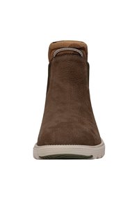 Botas de ante marrón hasta el tobillo con puntera redondeada, parte superior texturizada, cuello en contraste en tono beige y suela de goma blanca. Presenta paneles laterales elásticos.
