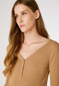 Lange mouwen, geribbelde top in bruin met een V-hals, voorzien van een knoopsluiting en een klein geborduurd logo aan de linkerkant.