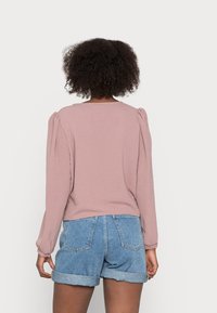 Femme aux cheveux bouclés portant un haut rose poussiéreux à manches longues et un short en denim bleu retroussé, debout de dos sur un fond blanc.