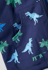 Tumesinine kangas koos siniste ja roheliste koomiksite dinosaurustega, sealhulgas T-Rex ja Triceratops, millel on õmmeldud patch tasku paremal küljel.