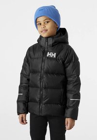 Dijete u crnoj puf jakni s kapuljačom i plavom pletenom kapom, stoji s opuštenim rukama naslonjeno na običnu bijelu pozadinu.