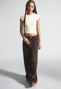 Beige cropped T-Shirt, enganzt geschnitten. Braune weit geschnittene Hose mit vorderem Knopf, seitlichen Taschen und Kordelzugdetails. Modell trägt braune Schuhe.