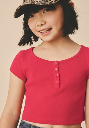 Jauna mergina su trumpais juodais plaukais dėvi leopardo rašto kepurę ir raudoną trumpomis rankovėmis susegamą palaidinę, lengvai šypsosi, fone šviesi aplinka.