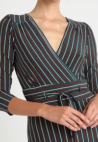 Zelené šaty s vertikálními pruhy v červené, bílé a tmavě zelené barvě. Maj�í hluboký V-výstřih, tříčtvrteční rukávy a detail s uvázaným páskem.