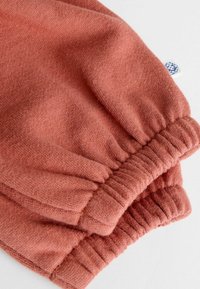 Nahaufnahme von staubig-pinker Stoff mit elastischen Bündchen und einem kleinen weißen Etikett, das ein blaues rundes Logo zeigt.