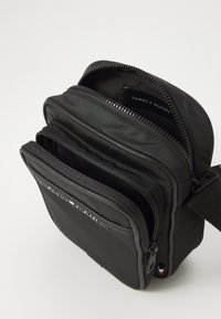 Geantă de umăr din nylon negru, cu multiple compartimente cu fermoar, având o curea robustă și un logo discret al mărcii în interior. Textură netedă.
