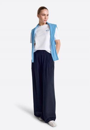 Frau steht, trägt ein weißes T-Shirt, einen hellblauen Pullover über den Schultern, weite marineblaue Hosen und weiße Slip-on-Schuhe.