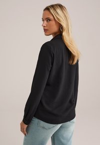 Donna con lunghi capelli biondi che indossa una maglietta nera a maniche lunghe e jeans azzurri chiari, che guarda oltre la spalla contro uno sfondo neutro.