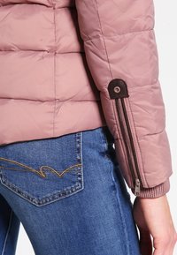 Różowa kurtka pikowana z dodatkowym ociepleniem i brązowymi akcentami zamka. Posiada ribrowane mankiety i dopasowany krój. Połączona z niebieskimi dżinsami.