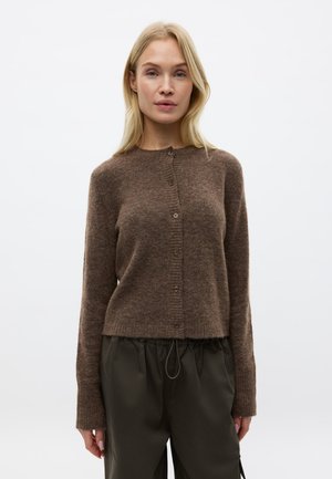 Femme blonde portant un cardigan marron boutonné et un pantalon vert foncé, debout devant un fond clair uni.