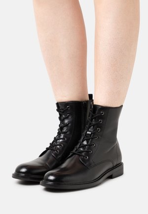 Botas de tobillo de cuero negro con un acabado suave, que cuentan con cordones y una cremallera lateral. Las suelas de goma planas proporcionan estabilidad.