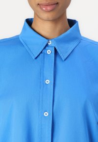 Camicia blu con colletto, caratterizzata da una trama liscia e cinque bottoni bianchi sulla parte anteriore. Design semplice e uniforme.