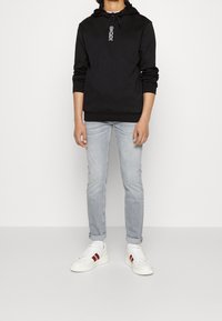 Sort hættebluse med en grafisk design på brystet, parret med lyseblå jeans og hvide sneakers med røde detaljer.