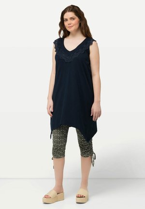 Marineblaue ärmellose Tunika mit Spitzenakzenten am Ausschnitt, kombiniert mit schwarzen gemusterten Capri-Leggings und beigen Plateausandalen.