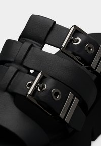 Gros plan sur des brides de sandales en cuir noir avec une surface texturée et des boucles et œillets en métal argenté brillant.