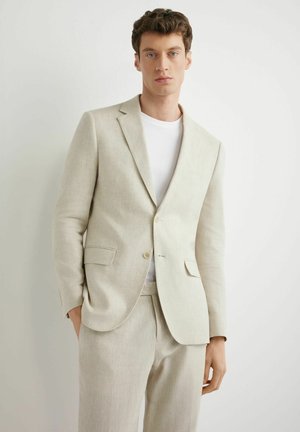 Młody mężczyzna z kręconymi włosami, ubrany w jasnobeżowy garnitur i białą koszulkę, stoi na jednolitym białym tle, ręce w kieszeniach.