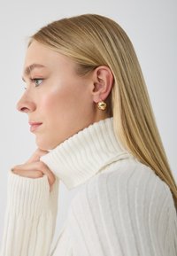 Pendientes esféricos de oro con un acabado pulido, llevados por una persona con un jersey de cuello alto blanco acanalado. Se destacan una textura suave y colores neutros.