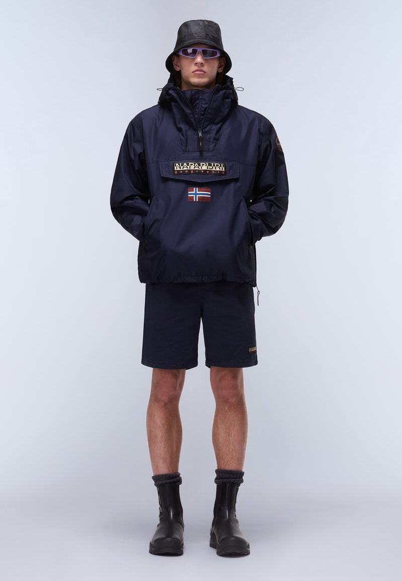 Marine windbreaker met half-rits, logopatch en vlagaccent, gecombineerd met bijpassende shorts en zwarte enkellaarzen, gedragen met een zwarte bucket hat.
