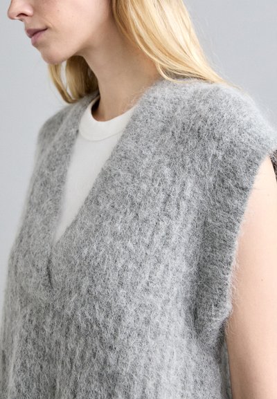 Szürke, texturált ujjatlan pulóver V-nyakkivágással, puha, bolyhos anyagból készült, látható varrással. Egy fehér ing fölött viselve.