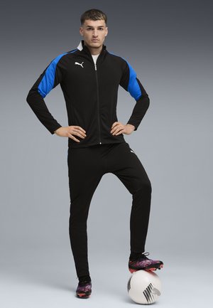 Zwarte sportieve trainingsoutfit met blauwe accenten, een gezoomde jas en aansluitende broek. Man staat op een voetbal, gekleed in kleurrijke voetbalschoenen.