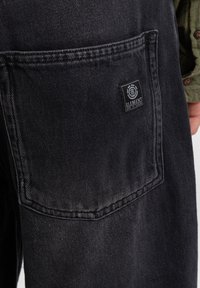 Pantalones de mezclilla negros con un bolsillo trasero que presenta una etiqueta en parche rectangular. La tela tiene una textura resistente y detalles de costura visibles.