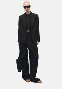 Blazer nero oversize con bottoni doppi, abbinato a pantaloni neri larghi e sandali piatti. Gli accessori includono occhiali da sole neri grandi.