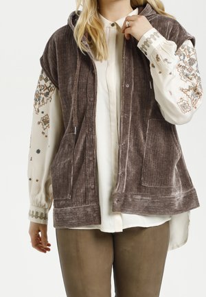 Personne portant une veste sans manches en velours côtelé marron par-dessus un chemisier blanc à manches longues avec des motifs floraux brodés sur les manches.