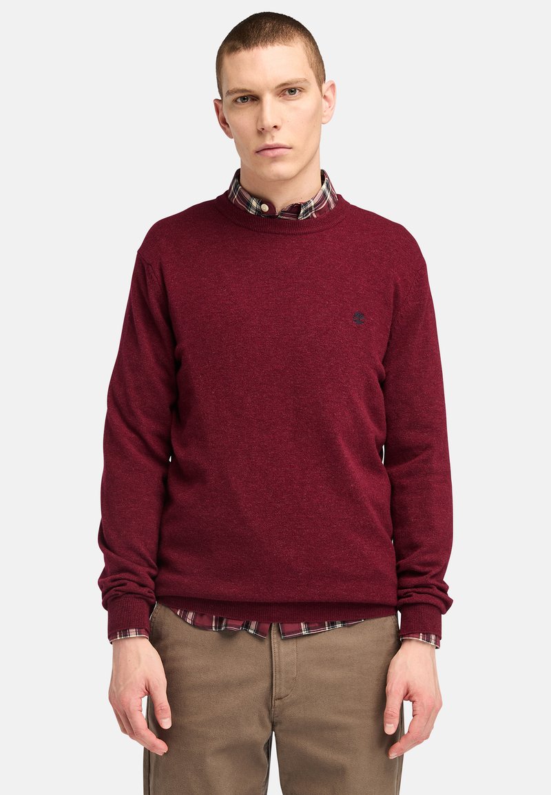 Burgundy rundhalsad tröja i stickad kvalitet, med ribbade ärmslut och nederkant, bärs över en rutiga skjorta och är matchad med beige byxor.