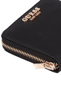Schwarze, strukturierte Brieftasche mit Reißverschluss an der Ecke, goldfarbenem Metallreißverschluss und goldfarbenem "GUESS Est. 1981"-Logo auf der Oberfläche.