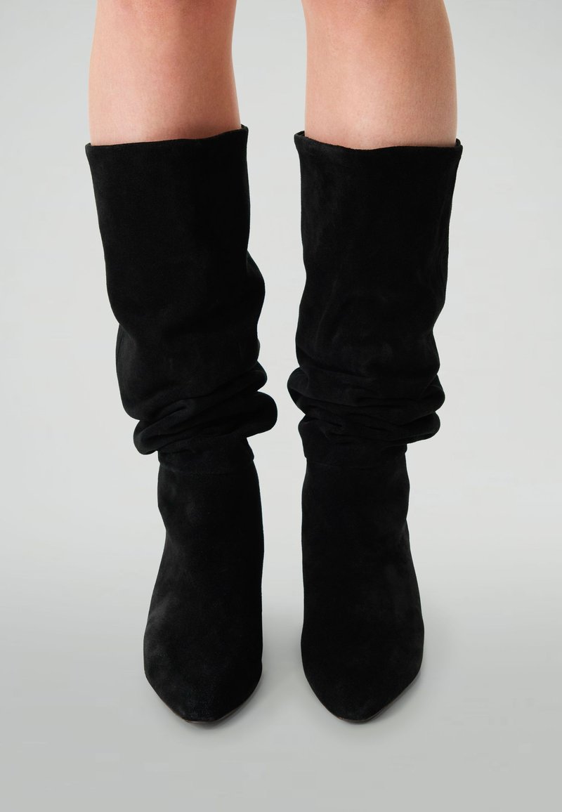 Femme portant des bottes en suède noires, montantes jusqu'aux genoux, à bouts pointus, se tenant sur une surface gris clair.