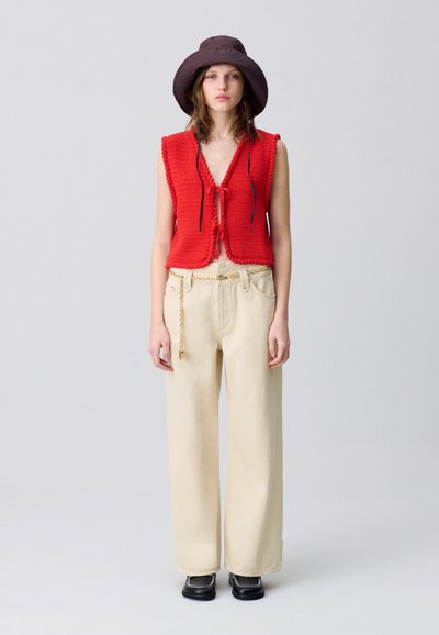 Chaleco de punto rojo con un diseño de lazo en la parte delantera, combinado con pantalones anchos beige y un sombrero marrón. Un calzado negro completa el look.