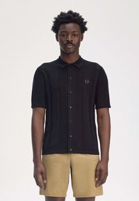 Polo shirt nero a maniche corte con texture a costine verticali, chiusura a bottoni e un logo ricamato discreto sul petto, indossato con pantaloni corti marroni.