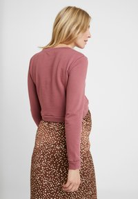 Top cropped de manga longa em tecido rosa suave, com decote redondo e punhos canelados, combinado com uma saia castanha com bolinhas creme.