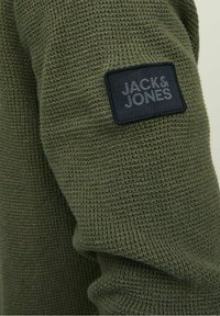Oliivinvihreä neulepaita, jossa on teksturoitu kudos. Sisältää mustan suorakulmaisen=laatan, jossa on painettuna "JACK & JONES".