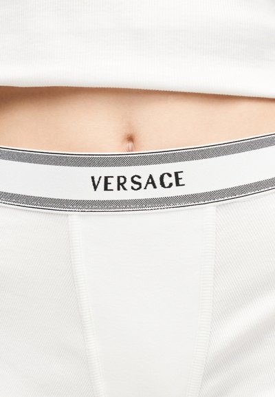 Gros plan sur la taille d'une personne portant un sous-vêtement blanc Versace avec une ceinture à logo et une chemise blanche couvrant partiellement le haut.