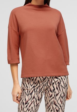 Mujer con una blusa de manga tres cuartos color óxido combinada con pantalones beige que tienen un estampado abstracto de animales en negro y rojo.