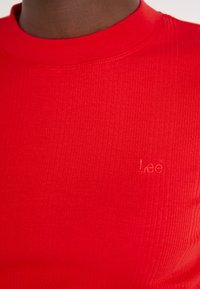 Crvena rebrasta majica s visokim izrezom i ušivenim "Lee" logotipom. Tkanina izgleda glatko i prilagođeno tijelu, ističući teksturu i vibrantnost boje.