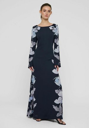 Donna che indossa un lungo abito blu navy con motivi floreali azzurri pallidi e grigi sulle maniche e sulla parte inferiore della gonna, in piedi davanti a uno sfondo semplice.