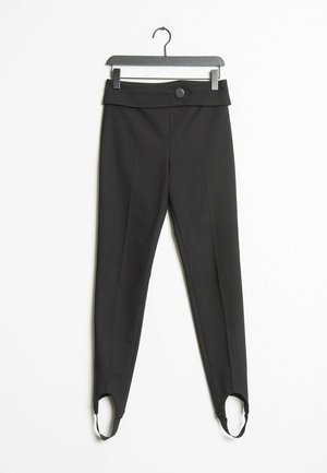 Schwarze, zulaufende Hose mit hohem Bund, vorderer Knopfleiste und Fußriemen, die an einem Bügel vor weißem Hintergrund hängen.