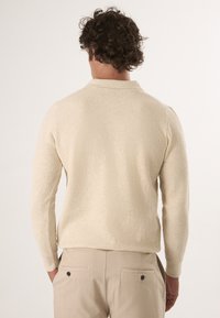 Beige gebreide pullover met textuurpatroon, lange mouwen en een klassieke kraag, gecombineerd met lichtbeige broeken met achterzakken.