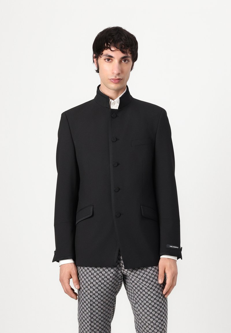 Black tailored blazer with a high collar, button closure, and side pockets. Smooth texture with contrasting details.